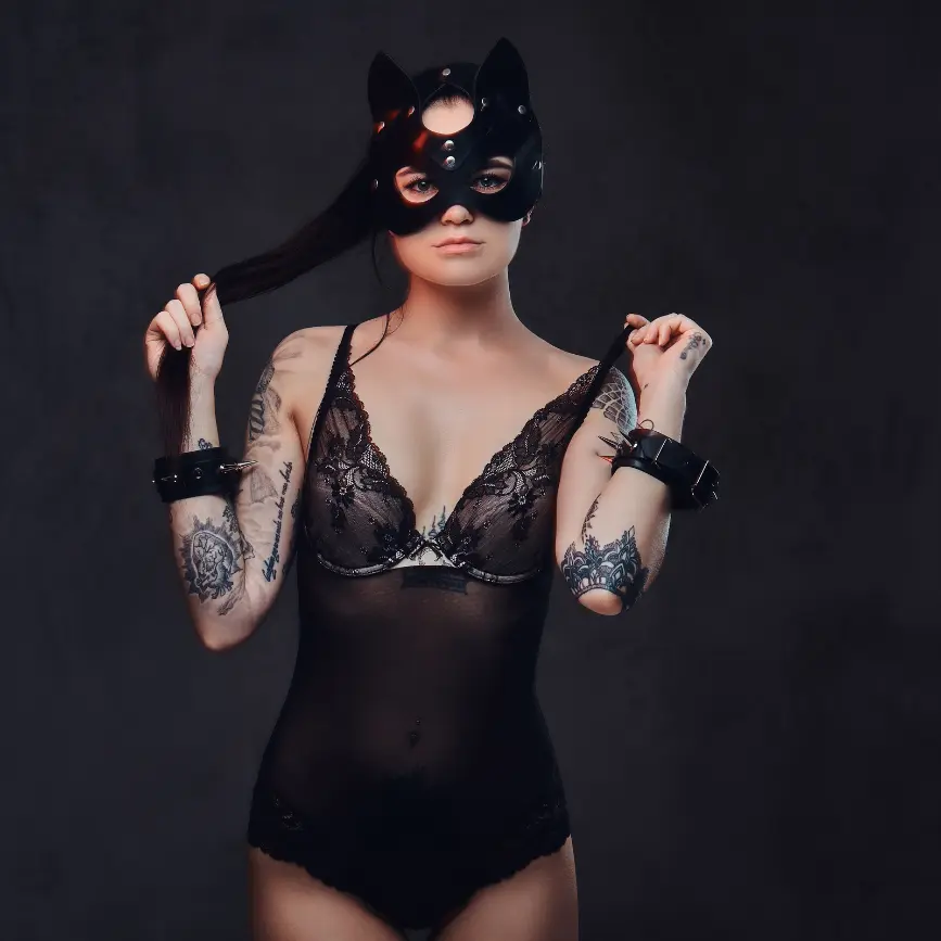 Mujer con máscara negra de gato, lencería de encaje y esposas de cuero, sosteniendo su cabello en un fondo oscuro.